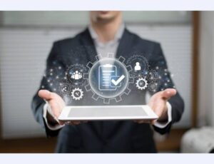 A businessman holds a tablet displaying digital icons of gears, checklists, and people, symbolizing productivity and teamwork.