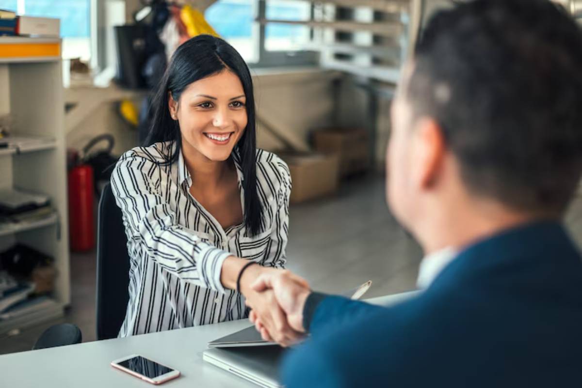 A woman in a striped shirt shakes hands with a man in a suit in a bright office setting, communicating professionalism and collaboration.