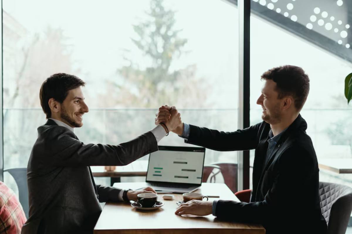 Two men in suits engage in a friendly arm wrestling match at a wooden table, with a laptop and coffee in a modern café setting.