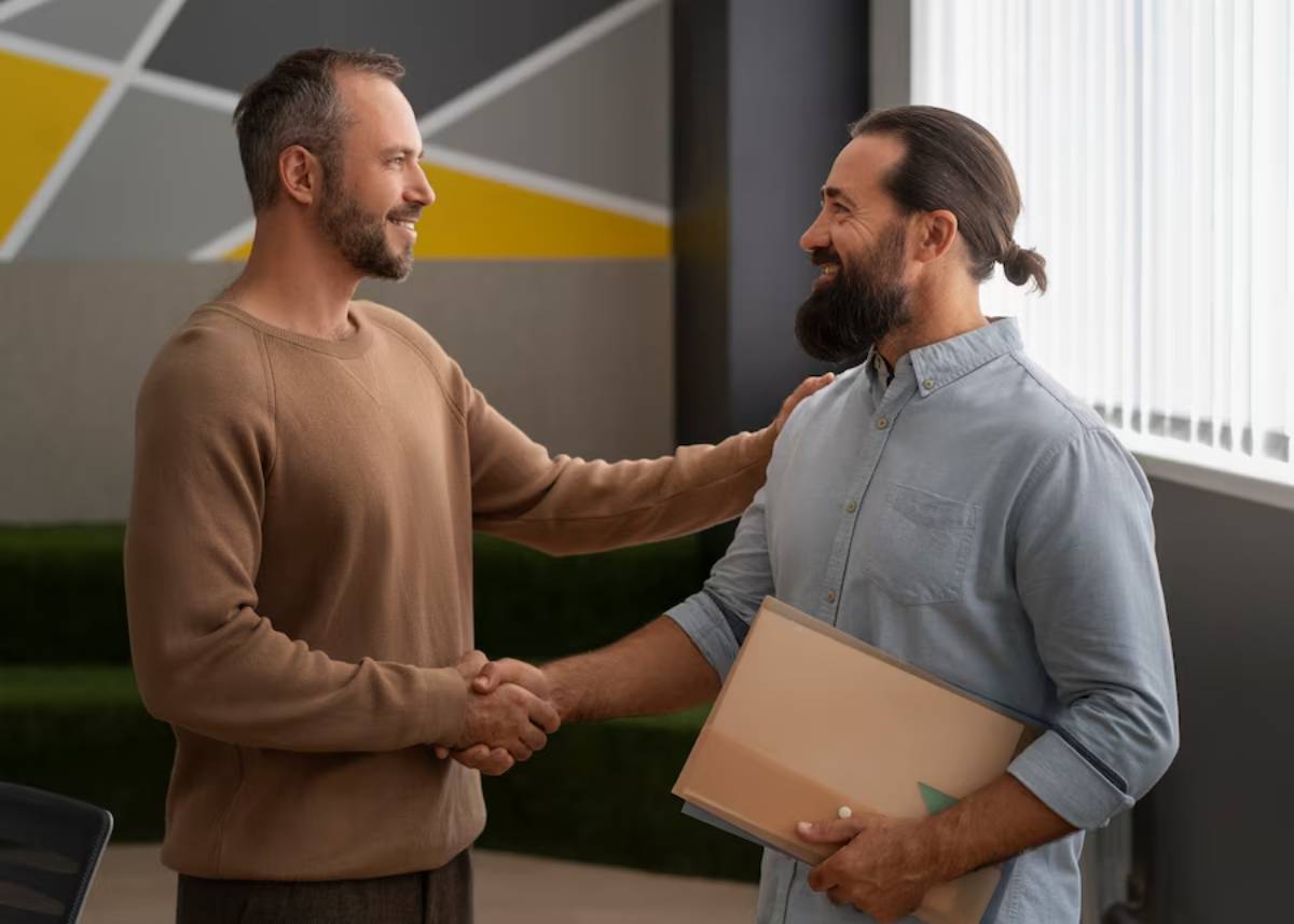 Two men engage in a handshake, one holding a folder, in a modern office with a geometric wall design.