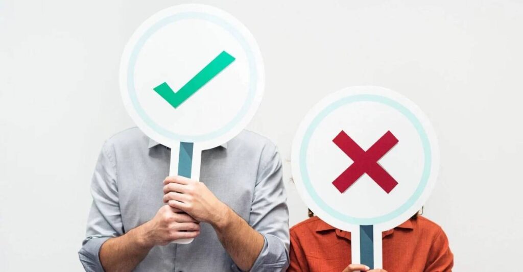 Two people hold large signs with a green checkmark and a red cross, symbolizing agreement and disagreement, in front of a plain background.