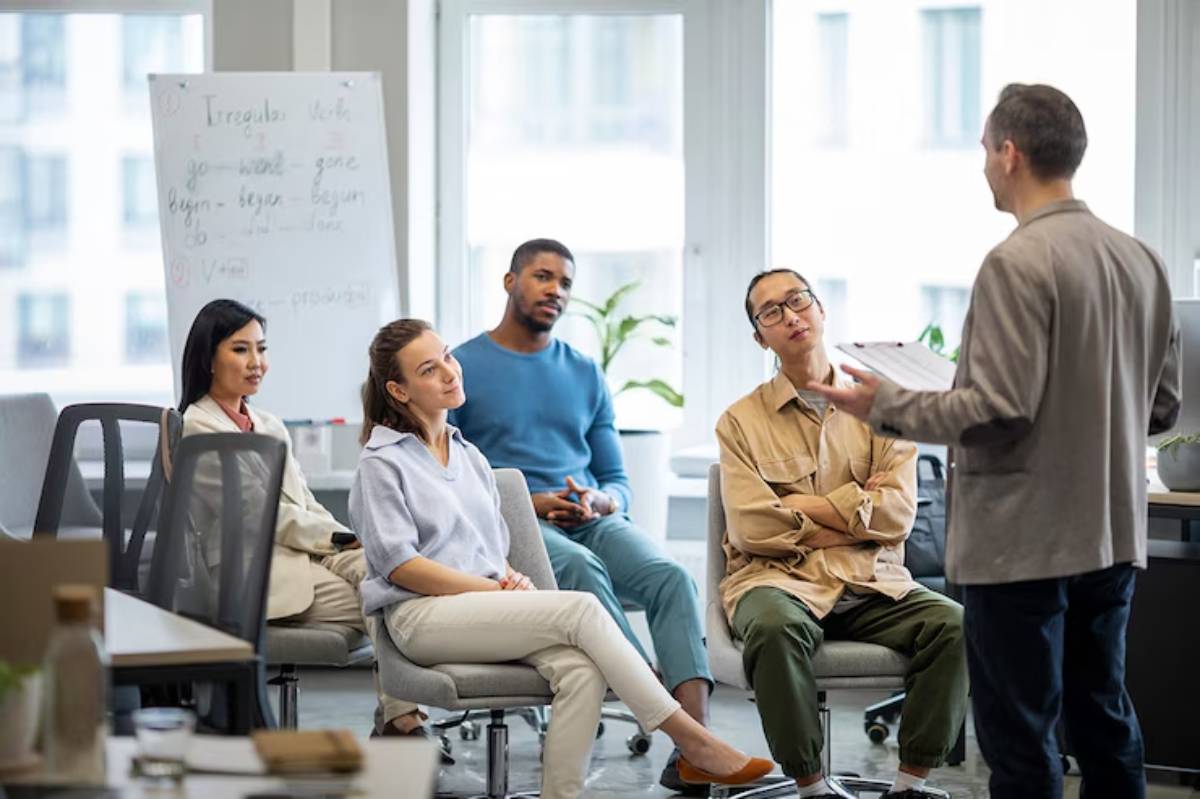 A group of diverse individuals seated in an office setting, attentively listening to a speaker presenting in front of a whiteboard.