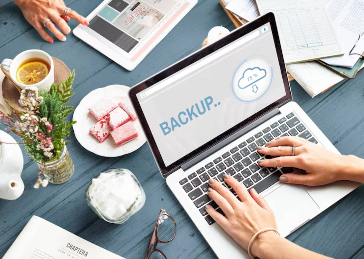 A laptop displaying a BACKUP screen at 75% progress, surrounded by a tea, sweets, flowers, and a tablet on a blue table.