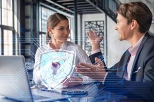 Two professionals engage in a discussion at a modern workspace, with a digital shield symbolizing cybersecurity overlaying the scene.
