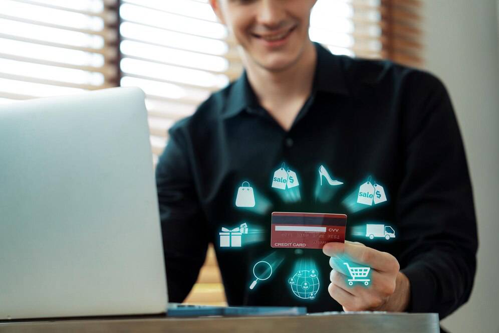 A person holds a credit card in one hand while shopping online, with various shopping icons glowing around a laptop.