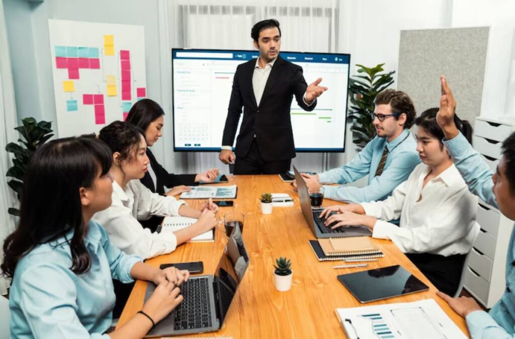 A business meeting with participants engaged at a conference table, laptops open, and a presenter addressing the group with a screen behind.