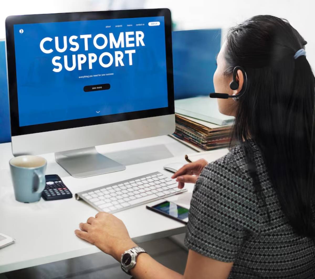 A woman in a headset works at a computer displaying a blue Customer Support webpage, with a coffee mug and calculator nearby.