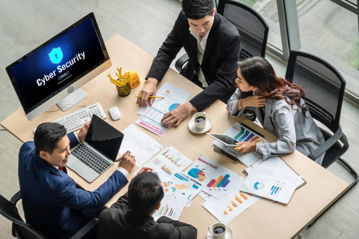 A group of professionals collaborates over charts and reports on cyber security at a modern conference table. A computer displays Cyber Security.