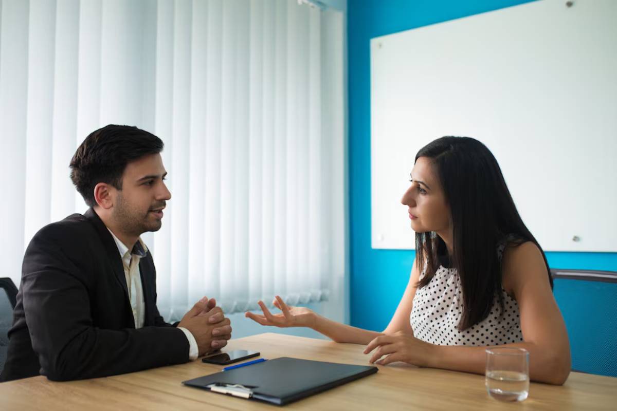 A man and woman engage in a discussion at a desk, with a notepad and glass of water visible; bright blue walls and curtains provide background.