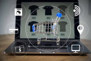 A mini shopping cart filled with cardboard boxes in front of a laptop displaying an online shopping site.