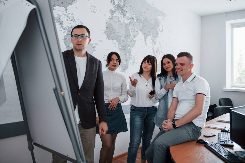A group of individuals stands in a bright office with a world map on the wall, engaged in discussion near a presentation board.