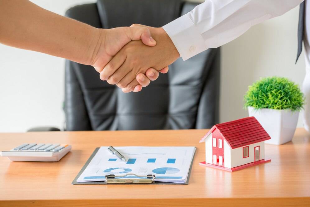 Two hands shaking over a desk with a small house model, charts, and a calculator, symbolizing a real estate agreement.