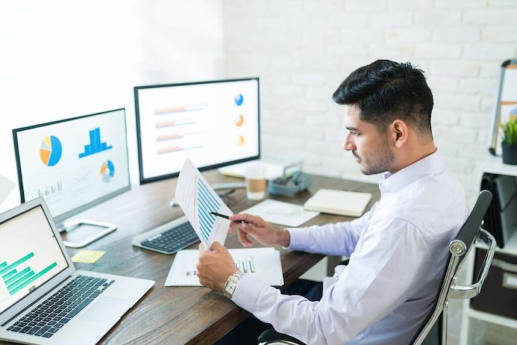 A man sits at a desk with multiple computer screens displaying graphs and charts, analyzing data while holding documents.
