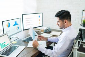 A man sits at a desk with multiple computer screens displaying graphs and charts, analyzing data while holding documents.