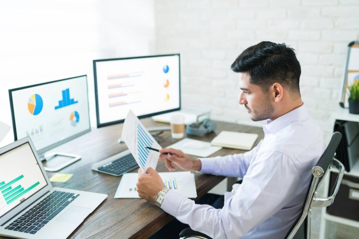 A man sits at a desk with multiple computer screens displaying graphs and charts, analyzing data while holding documents.