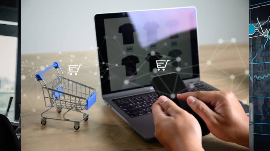 A person holds a smartphone while shopping online on a laptop, with a miniature shopping cart visible on the table.
