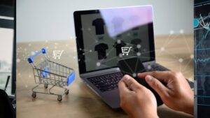 A person holds a smartphone while shopping online on a laptop, with a miniature shopping cart visible on the table.