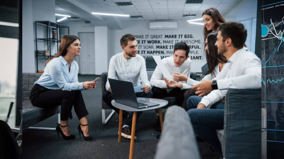 A group of professionals collaboratively discussing ideas around a laptop in a modern office setting with motivational wall text.
