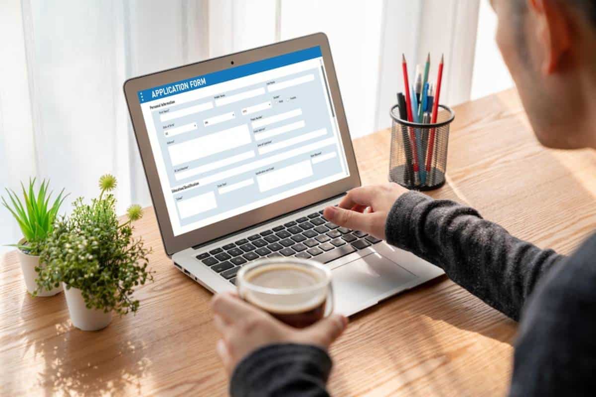 A person fills out an application form on a laptop while holding a cup of coffee, with plants and stationery in the background.