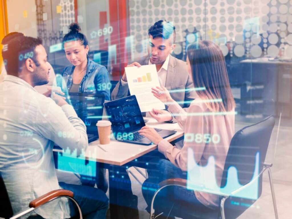 A group of professionals collaborate at a table, analyzing data on laptops while reviewing a report with graphics and statistics.
