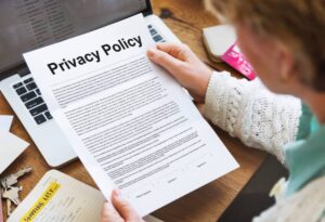 An older adult examines a printed Privacy Policy document while seated at a desk with a laptop and a notepad.