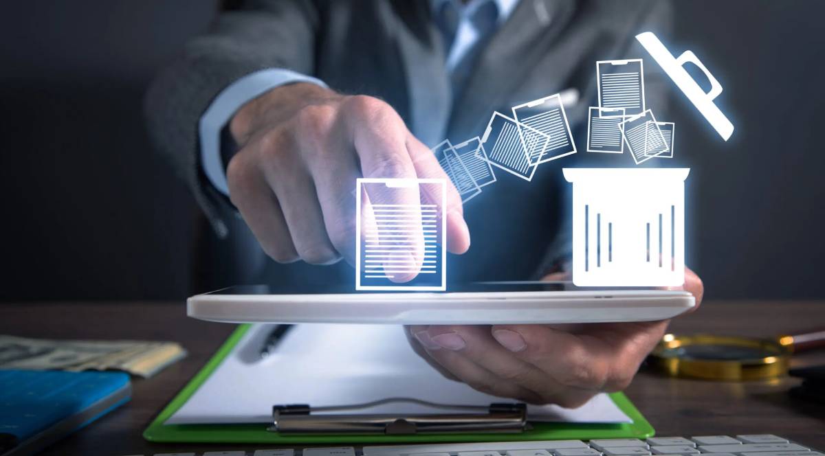 A businessperson's hand gestures towards a digital tablet displaying documents being deleted into a trash bin icon.
